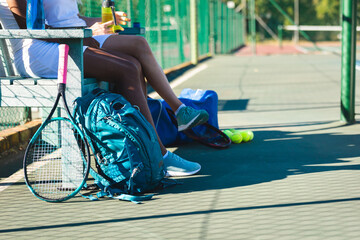 Low section of multiracial female players sitting on bench at tennis court during break on sunny day - Powered by Adobe