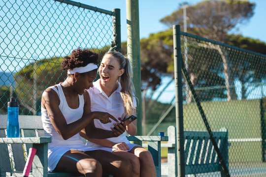 Cheerful biracial female tennis players sharing smartphone while sitting on bench at court