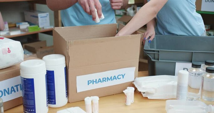 Close up of males and females volunteers putting medicines inside cardboard box while working together at charitable foundation. Social workers preparing packages with goods for first aid.