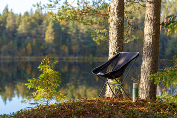 Beautiful forest lake with camping folding chair, book and vacuum insulated bottle.