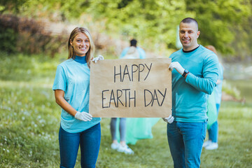 Diverse Group of People Picking Up Trash in The Park Volunteer Community Service. Happy international volunteers holding placard with 'happy Earth day' message.