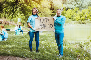 Diverse Group of People Picking Up Trash in The Park Volunteer Community Service. Happy international volunteers holding placard with 'happy Earth day' message.