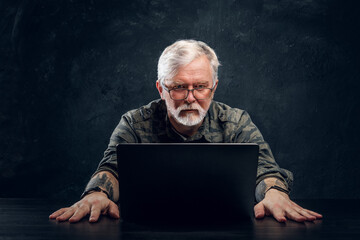 Photo of contemporary old man with tattoo sitting at table there is laptop against dark background.