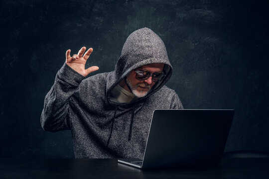 Studio shot of tricky elderly hacker sitting at table with laptop against dark background.