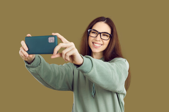 Student girl takes selfie standing against solid brown studio background. Happy young woman with long hair and toothy smile in hoodie and glasses takes picture of herself on her mobile phone
