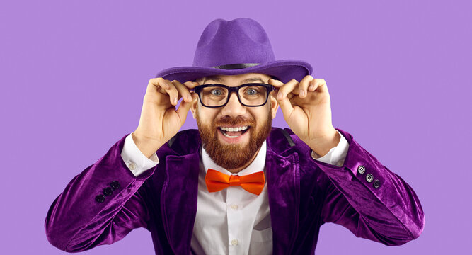 Portrait of funny cheerful stylish showman who wears glasses while standing on purple background. Caucasian bearded man in stylish purple jacket and hat is smiling madly looking at camera. Banner.