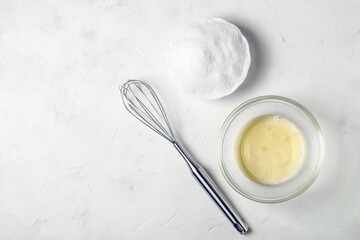 ingredients for the French meringue dessert are egg white, sugar and a whisk on a gray surface, copy space.