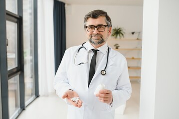 Senior male doctor with stethoscope in medical scrubs and holding bottle and tablets