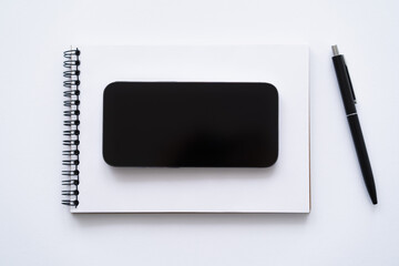 top view of smartphone with blank screen on notebook near pen on white.