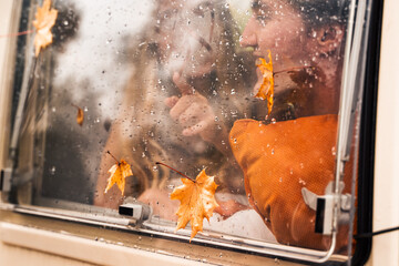 Young happy lovers couple behind wet misted window,rain drops.Drawing heart with finger.Autumn atmosphere,mood,maple leaves.Love romantic road travel. Traveling in camper,house on wheels,trailer