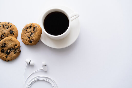 top view of cup of coffee near chocolate chip cookies and wired earphones on white.
