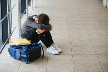 Little boy sitting alone on floor after suffering an act of bullying while children run in the...
