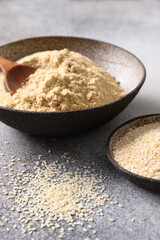 Sesame flour in bowl and seeds for clean eating gluten-free dessert and pastry on gray background. Vertical. Close up.