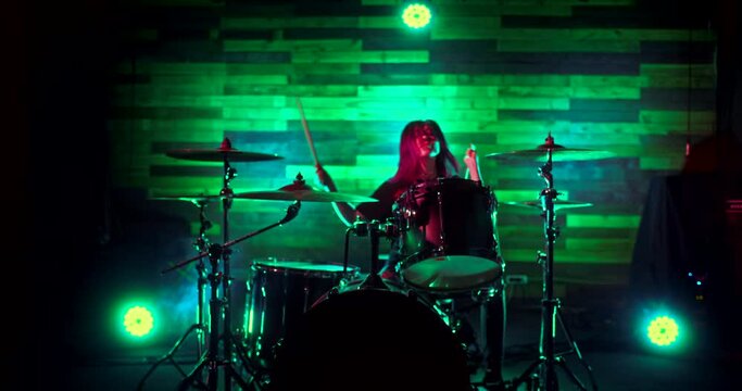 Young beautiful girl playing drums on the stage of nightclub. Light show during the concert. The light flickers to the beat of the drums. Camera moves away from girl drummer