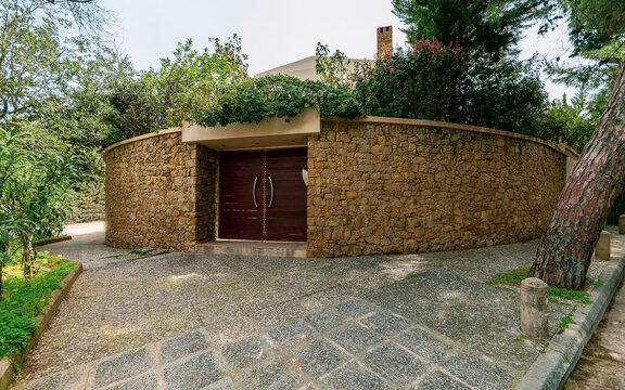 A house behind a tall stone wall fence with a natural dark brown wood door, Athens, Greece.