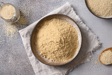 Sesame flour in bowl and white sesame seeds on gray background for cooking low carbohydrate dessert. Top view.