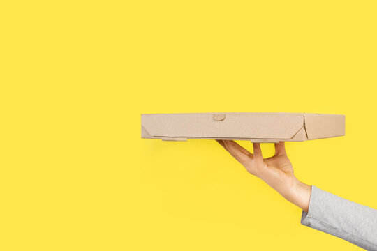 Mano de mujer sosteniendo una caja de cart&oacute;n marr&oacute;n ecol&oacute;gica de pizza sobre un fondo amarillo brillante liso y aislado. Vista de frente y de cerca. Copy space