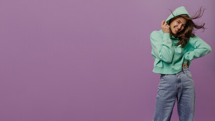 Happy young caucasian girl with tousled hair in hood looks at camera on purple background. Brunette smiles and wears hoodie with jeans. Good mood concept