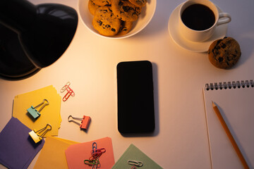 top view of lamp near smartphone with blank screen, chocolate chip cookies, cup of coffee and stationery on white.