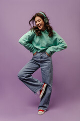 Full length happy young caucasian woman spends leisure time listening to favorite song through headphones in studio. Brunette wear casual clothes keeps hands in pockets and looks down at her feet.