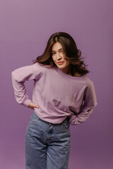 Cute young caucasian girl with dark wavy hair looks at camera on purple background. Brunette in sweater and jeans keeps her hands on waist. Lifestyle concept