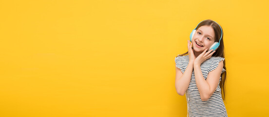 happy little girl with dark hair listens to music in headphones and looks at camera smiling on yellow background, space for text, banner