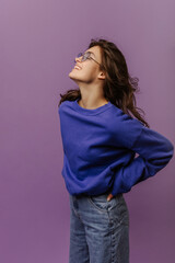 Cute young caucasian girl in glasses closing eyes smiles on purple background. Brunette wearing sweatshirt and jeans keeps her hands on hips. Lifestyle, different emotions, leisure concept.