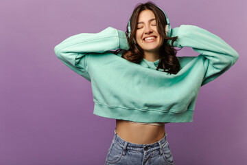 Happy young caucasian girl closing eyes smiling holding headphones with hands on purple background. Brunette woman with wavy hair wears hoodie and jeans. Lifestyle, emotions, leisure concept.