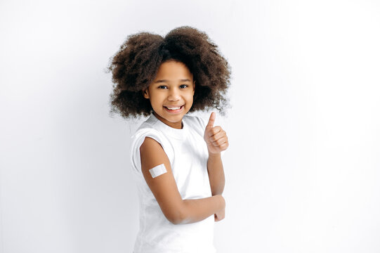 Positive African American little girl, preschooler, with band-aid on shoulder after vaccination, for immunity against covid19, and other diseases, stand on isolated white background, thumb-up gesture