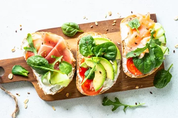 Zelfklevend Fotobehang Open sandwichset met roomkaas, prosciutto, zalm, avocado en verse groenten. Bovenaanzicht aan witte tafel. © nadianb
