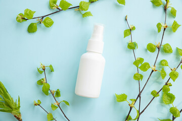 White cosmetic spray bottle and birch branches with young small leaves on blue background. Mockup...