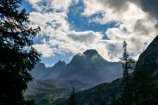 
Eine majest&auml;tische Bergspitze ragt unter einem dramatisch bew&ouml;lkten Himmel empor, eingerahmt von dunklen Nadelb&auml;umen im Vordergrund einer alpinen Landschaft.