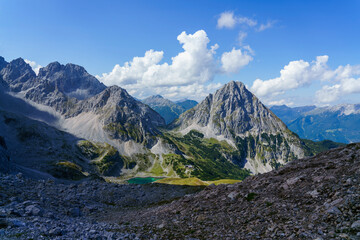 Eine weite Berglandschaft unter blauem Himmel mit weißen Wolken offenbart felsige Gipfel, grüne...