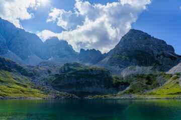 Ein glasklarer Bergsee spiegelt die imposanten, teils wolkenverhangenen Gipfel einer schroffen Alpenlandschaft unter strahlend blauem Himmel wider.