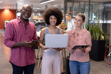Portrait of smiling multiracial colleagues standing with wireless technologies at modern workplace