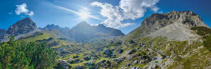 Obraz premium Ein weites Panorama zeigt eine beeindruckende Gebirgskette unter strahlend blauem Himmel, während die Sonne hell über den schroffen, teils begrünten Felsgipfeln und einem steinigen Tal leuchtet.