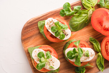 Top view on bruschettas with caprese salad, sliced tomatoes, mozzarella, basil and topped with corn salad on wooden serving board, white table background with copy space. Classic italian starter