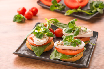 Bruschettas with caprese salad, sliced tomatoes, mozzarella, basil and topped with corn salad on black plate, wooden table background. Classic italian starter