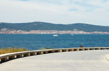 Fototapeta na wymiar Passing a paved road laid along sea coast. A boat sails in distance against the backdrop of a coastal resort