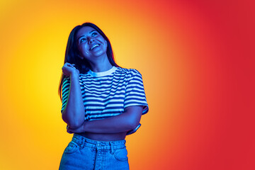 Portrait of young girl dreamly smiling, posing isolated over red yellow background in neon light