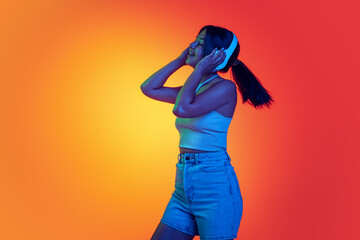 Young cheerful woman listening to music in headphones isolated over gradient yellow orange background in neon light