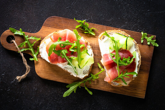 Open sandwich with cream cheese and prosciutto or jamon with fresh greens. Top view at dark.