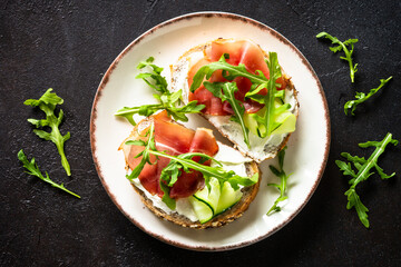 Open sandwich with cream cheese and prosciutto or jamon with fresh greens in white plate. Top view at dark table.