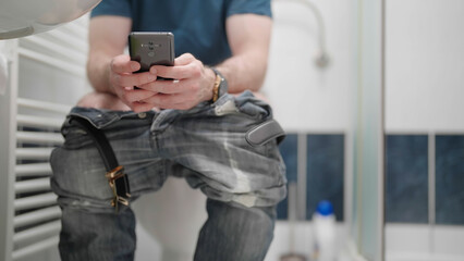 Headless male person in bathroom browsing on smartphone while on toilet - obrazy, fototapety, plakaty
