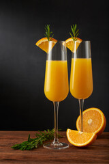 A pair of mimosa cocktail in flute glass with orange slice and rosemary twig on a wooden bar and dark background.