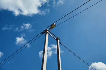 A street lamp hangs on concrete poles, black electric wires go to the poles, a blue sky with clouds, a view from below, a simple street composition