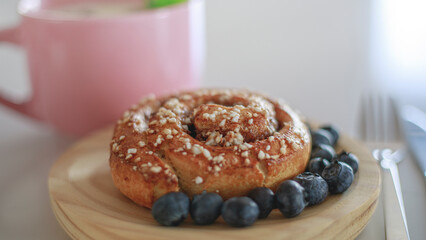 Close-up of a cinnamon roll and blueberries around it on a wooden plate with a fork next to it in a bright, light-filled setting