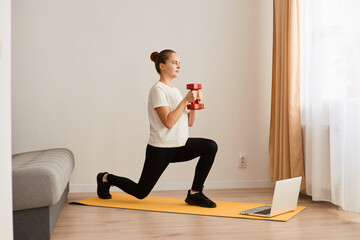 Obraz premium Indoor shot of young athletic sporty woman training her body at home, using laptop for watching tutorial sports videos, holding dumbbells and doing lunges.