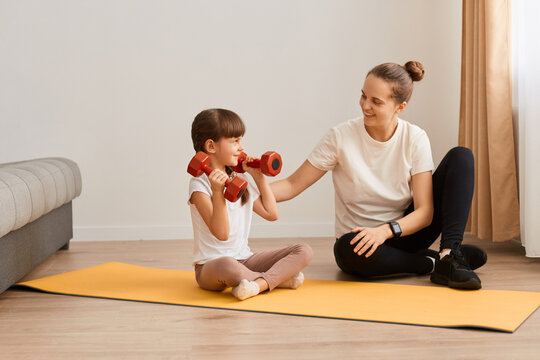 Image of woman trainer having workout with with little girl wearing sportswear doing sport exercises for arms with dumbbells, sitting on floor, doing exercise for biceps and triceps.