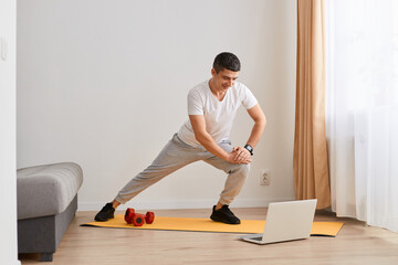 Fit gymnast man with dark hair in sportswear standing on mat and stretching legs, warming up, doing sports flexibility exercises, fitness and yoga workout at home.